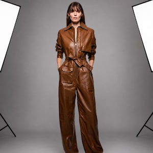 May include: A brown leather jumpsuit featuring a collared neckline, a full-length front zipper, and long sleeves. The garment includes a tie-waist belt, two front flap pockets at the hip, and wide-leg trousers that extend to the floor.