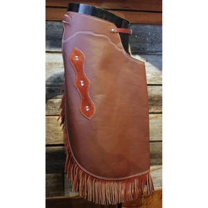 May include: Brown leather chaps with decorative stitching and a fringed bottom edge. Features an orange-brown leather accent with three silver rivets. A red leather strap is visible near the top.