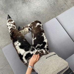 Peut inclure: Pantalon &agrave; motif peau de vache avec des talons noirs &agrave; bout pointu. Le pantalon pr&eacute;sente un m&eacute;lange de taches marron, noires et blanches. Un sac gris clair avec une fermeture &eacute;clair noire et la marque "PRADA" est &eacute;galement visible.