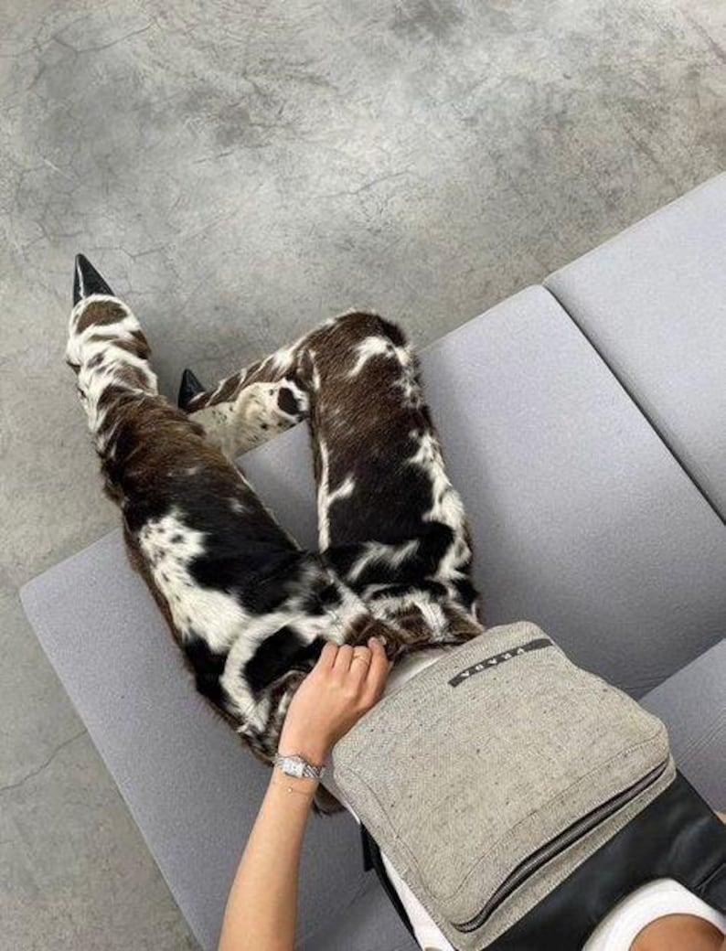 Peut inclure: Pantalon &agrave; motif peau de vache avec des talons noirs &agrave; bout pointu. Le pantalon pr&eacute;sente un m&eacute;lange de taches marron, noires et blanches. Un sac gris clair avec une fermeture &eacute;clair noire et la marque "PRADA" est &eacute;galement visible.