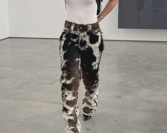 May include: A person wearing a white tank top, a black leather harness, and cow-print pants. The pants are brown, black, and white with a fur-like texture. The shoes are black with pointed toes.