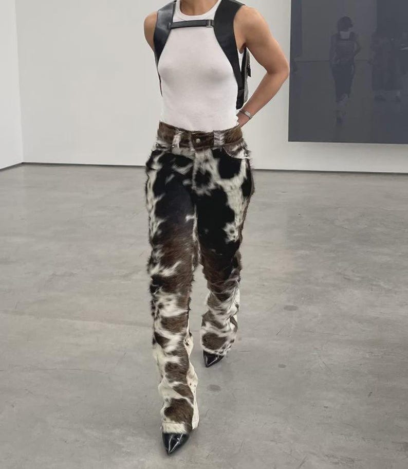 Peut inclure: Une personne porte un d&eacute;bardeur blanc, un harnais en cuir noir et un pantalon &agrave; imprim&eacute; vache. Le pantalon est marron, noir et blanc avec une texture semblable &agrave; de la fourrure. Les chaussures sont noires avec des bouts pointus.