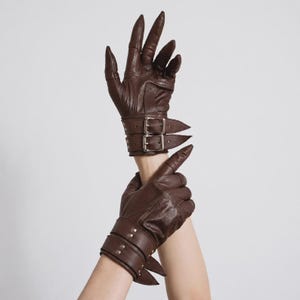 May include: A pair of dark brown leather gloves featuring pointed, claw-like fingertips and two adjustable wrist straps with silver-toned metal buckles. The gloves have a smooth finish with visible stitching and reinforced panels across the knuckles.