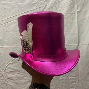Pode incluir: Um chap&eacute;u alto magenta com acabamento met&aacute;lico. O chap&eacute;u apresenta uma faixa decorativa e &eacute; adornado com penas brancas e salpicadas. O chap&eacute;u &eacute; segurado por uma pessoa.