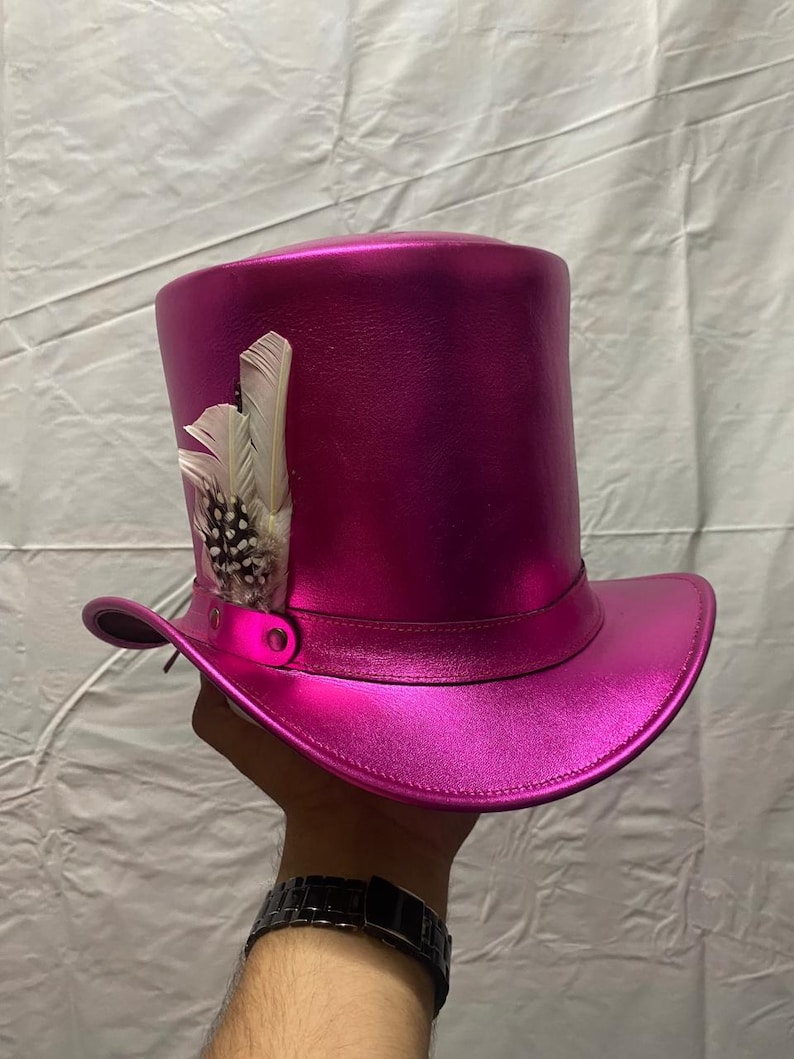 Pode incluir: Um chap&eacute;u alto magenta com acabamento met&aacute;lico. O chap&eacute;u apresenta uma faixa decorativa e &eacute; adornado com penas brancas e salpicadas. O chap&eacute;u &eacute; segurado por uma pessoa.