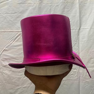 Pode incluir: Um chap&eacute;u alto magenta com um acabamento brilhante e met&aacute;lico. O chap&eacute;u tem uma aba larga e uma coroa alta e cil&iacute;ndrica. Um pequeno bot&atilde;o decorativo est&aacute; na lateral da aba. O chap&eacute;u &eacute; segurado por uma m&atilde;o.