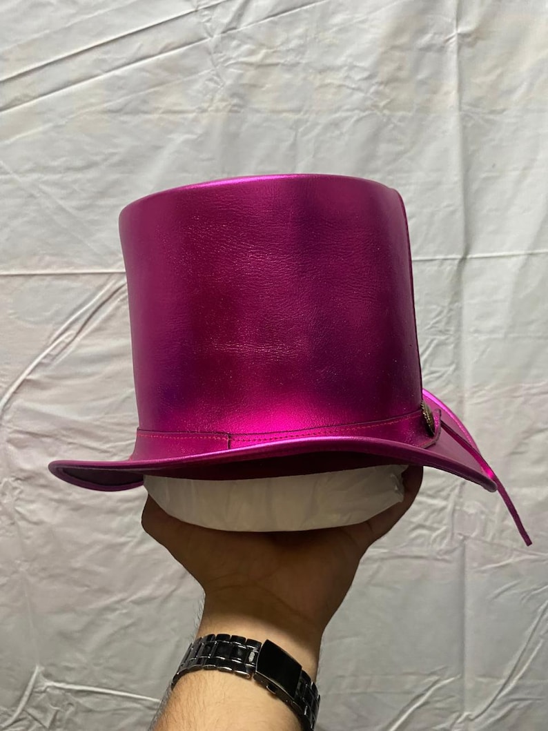 Pode incluir: Um chap&eacute;u alto magenta com um acabamento brilhante e met&aacute;lico. O chap&eacute;u tem uma aba larga e uma coroa alta e cil&iacute;ndrica. Um pequeno bot&atilde;o decorativo est&aacute; na lateral da aba. O chap&eacute;u &eacute; segurado por uma m&atilde;o.