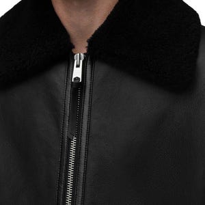 K&ouml;nnte beinhalten: Schwarze Lederjacke mit schwarzem Shearling-Kragen. Die Jacke hat einen silbernen Rei&szlig;verschluss und eine strukturierte Lederoberfl&auml;che. Das Bild ist eine Nahaufnahme der Jacke, die sich auf den Kragen und den Rei&szlig;verschluss konzentriert.