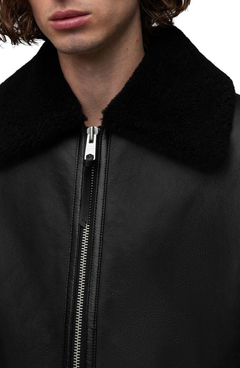 K&ouml;nnte beinhalten: Schwarze Lederjacke mit schwarzem Shearling-Kragen. Die Jacke hat einen silbernen Rei&szlig;verschluss und eine strukturierte Lederoberfl&auml;che. Das Bild ist eine Nahaufnahme der Jacke, die sich auf den Kragen und den Rei&szlig;verschluss konzentriert.