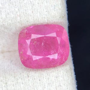 Puede incluir: Una gema facetada, de talla cojín, en un vibrante tono rosa. La gema se encuentra sobre un fondo de tela blanca, con un telón de fondo oscuro. El corte de la piedra revela múltiples facetas, realzando su brillo.