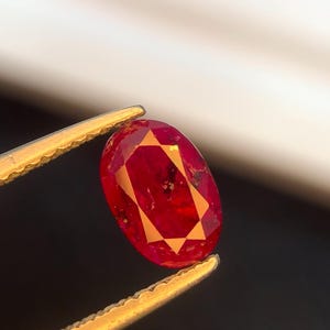 May include: A faceted, oval-shaped ruby gemstone held by gold tweezers. The ruby is a deep red color with lighter orange-red facets. The gemstone has a few dark inclusions. The tweezers are in focus, and the background is blurred.