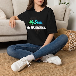 Może przedstawiać: Czarny t-shirt z krótkim rękawem z napisem My Data w zielono-niebieskim gradiencie nad napisem MY BUSINESS białymi wielkimi literami, z subtelnym ciemnym kolistym wzorem za tekstem, w połączeniu z niebieskimi dżinsami i białymi trampkami.