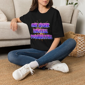 Może przedstawiać: Czarny t-shirt z krótkim rękawem z nadrukiem graficznym przedstawiającym złotą iskrę oraz napis "ONE SPARK ENDLESS POSSIBILITIES" w fioletowo-niebieskim gradiencie, zestawiony z ciemnoniebieskimi dżinsami i białymi trampkami.
