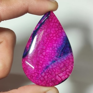 Puede incluir: Una gema en forma de lágrima con un vibrante patrón de color rosa y azul. La superficie pulida muestra un efecto texturizado y agrietado, con vetas azules más oscuras que atraviesan la base rosa. La gema se sostiene entre dos dedos.