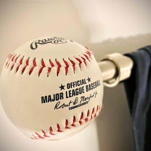 May include: A white leather baseball with red stitching mounted on a silver metal rod. The ball features the Rawlings logo and the text Official Major League Baseball Robert D. Manfred Jr. Commissioner printed in black ink on the surface.