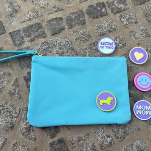 Könnte beinhalten: Eine türkisfarbene Clutch mit Handschlaufe und goldenem Reißverschluss. Die Clutch hat eine Dackel-Hunde-Anstecknadel und weitere Anstecknadeln mit einem Herz, einem Smiley und den Worten "MOM OF TWO" und "MOM WOW".