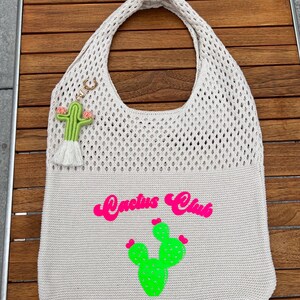 Könnte beinhalten: Eine beige Stricktasche mit einem oberen Netzbereich und einem festen unteren Teil. Die Tasche zeigt eine leuchtend pinke Grafik mit der Aufschrift "Cactus Club" über einer grünen Kaktus-Illustration. Ein kleiner grüner Kaktus-Schlüsselanhänger ist befestigt.