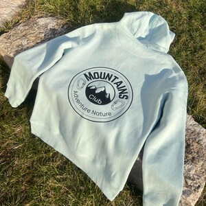 Könnte beinhalten: Ein hellblauer Kapuzenpullover mit einem schwarzen Kreislogo auf der Rückseite, auf dem "Mountains Club Adventure Nature" steht.