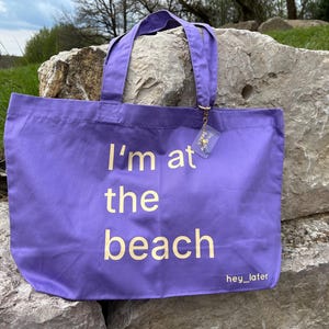 Könnte beinhalten: Eine lila Tragetasche mit dem Text "I'm at the beach" in weißer Schrift. Die Tasche hat passende lila Griffe und ein kleines Etikett. Die Worte "hey_later" sind am unteren Rand der Tasche aufgedruckt.
