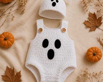 May include: A white crocheted baby romper and matching beanie set featuring a ghost face design. The romper has two brown wooden buttons at the shoulder straps and three black felt-like ovals forming eyes and a mouth. The beanie has a small knot.