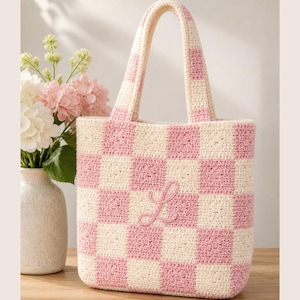 May include: A handmade crochet tote bag with a pink and cream checkerboard pattern. The bag features a matching handle and a cursive "L" embroidered on the front. The bag is displayed next to a vase of pink and white flowers.