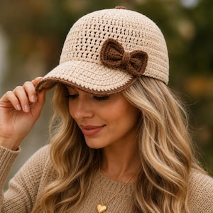 May include: A beige crochet baseball cap featuring a dark brown crocheted bow attached to the side. The cap has a structured crown with a textured stitch pattern and a dark brown trim along the edge of the curved brim, creating a two-tone appearance.