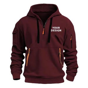 May include: A maroon hoodie with a half-zip closure, a hood, and a kangaroo pocket. The hoodie features a small pocket on the left sleeve and a zippered pocket on the right sleeve. The text "YOUR DESIGN" is printed on the chest.
