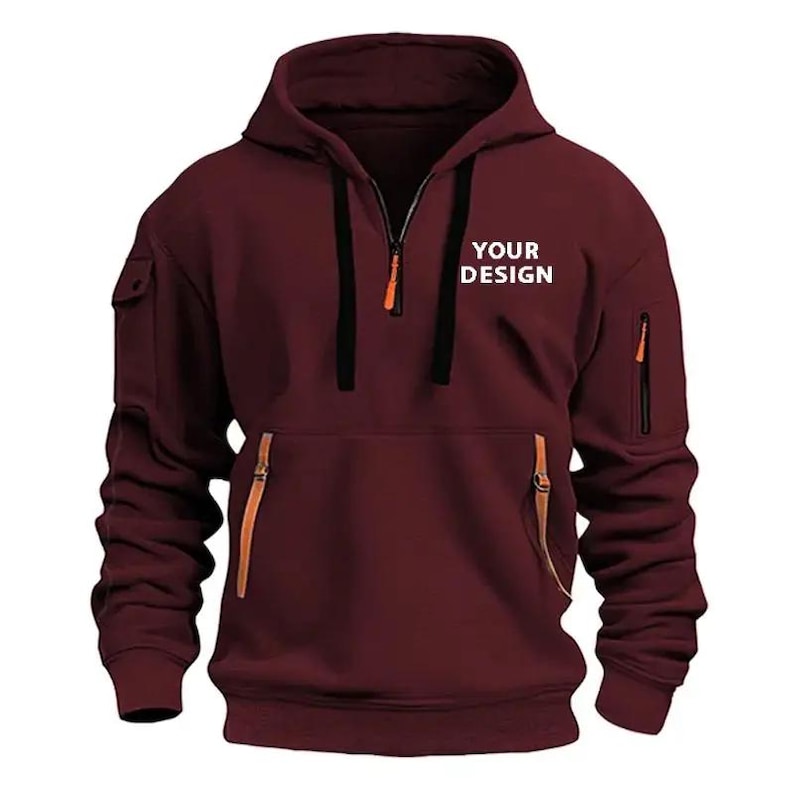 May include: A maroon hoodie with a half-zip closure, a hood, and a kangaroo pocket. The hoodie features a small pocket on the left sleeve and a zippered pocket on the right sleeve. The text "YOUR DESIGN" is printed on the chest.