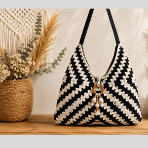 May include: A black and white crochet shoulder bag with a geometric pattern. The bag has a wooden ring and beaded tassel detail. The bag is sitting on a wooden surface.
