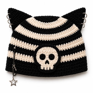 May include: A black and cream striped crochet beanie featuring cat-ear points with three silver metal rings on each side. A circular patch with a cream skull on a black background is centered on the front, and a small silver star charm hangs from a chain.