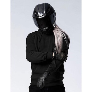 May include: A black motorcycle helmet with a dark visor and a carbon fibre weave pattern. The person is wearing a black hoodie and black leather gloves. Long, silver hair is visible.