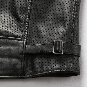 May include: Close-up of a black leather jacket with a perforated design. The jacket features a buckle closure on the sleeve and quilted detailing. The leather appears to be high-quality, with a matte finish.
