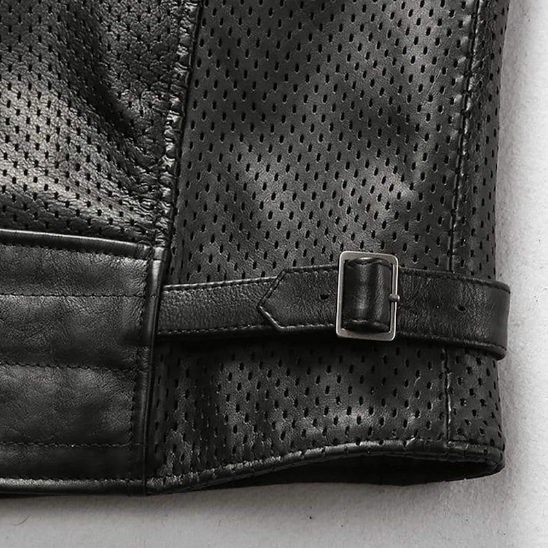 May include: Close-up of a black leather jacket with a perforated design. The jacket features a buckle closure on the sleeve and quilted detailing. The leather appears to be high-quality, with a matte finish.