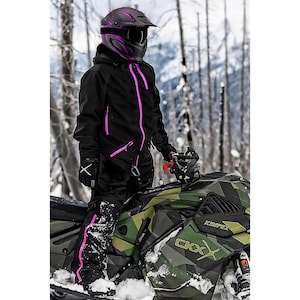 Może przedstawiać: Czarny kombinezon na skuter śnieżny z jasnoróżowymi zamkami i akcentami, w połączeniu z pasującym kaskiem. Kask ma wizjer i różowe detale. Skuter śnieżny jest w zielono-brązowym kamuflażu.
