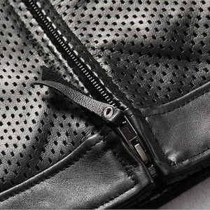 May include: Close-up of a black leather jacket with a perforated design. The jacket features a zipper closure and a leather pull tab. The leather has a matte finish and the stitching is visible.