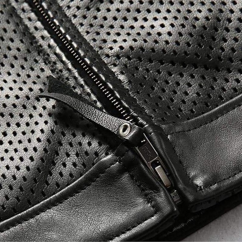 May include: Close-up of a black leather jacket with a perforated design. The jacket features a zipper closure and a leather pull tab. The leather has a matte finish and the stitching is visible.
