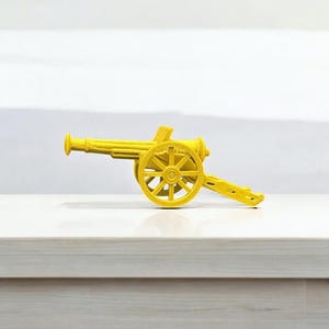May include: A small, yellow toy cannon with a detailed design. The cannon features a barrel, wheels, and a carriage, all in a bright yellow color. The toy is set against a white background, highlighting its form.
