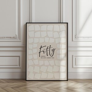 May include: A framed print with a beige background and a black border. The print features the word "Fifty" in elegant script, with the phrase "Things We Love About You" below. The background is filled with off-white, square shapes.