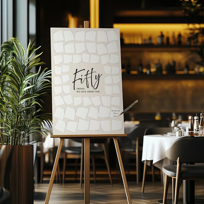 May include: A wooden easel displays a sign with the word "Fifty" in elegant script, followed by "Things We Love About You." The sign has a light beige background with white square shapes. A pen is poised to write on the sign.