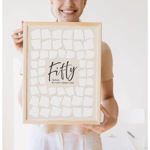 May include: A framed print with the word "Fifty" in elegant script, with the text "THINGS WE LOVE ABOUT YOU" below. The print features a grid of blank, off-white shapes on a light beige background, perfect for personalized notes.