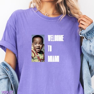 May include: A lilac t-shirt with the words "WELCOME TO MIAMI" in white. It features a photo of a young boy. The shirt has short sleeves and a relaxed fit.