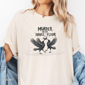 Könnte beinhalten: Elfenbeinfarbenes T-Shirt mit dem Text "MURDER ON THE DANCE FLOOR". Die Grafik zeigt zwei Krähen mit ausgebreiteten Flügeln, die sich gegenüberstehen, mit einer Discokugel dazwischen. Das Design ist in Schwarz und Weiß gehalten.
