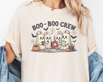 May include: Ivory t-shirt with a Halloween graphic. The design shows cartoon ghosts in nurse uniforms, with the text "BOO-BOO CREW". Bats, spiderwebs, and pumpkins are also featured in the illustration.