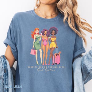 以下が含まれることがあります： スタイリッシュな服装の女性3人のグラフィックがプリントされた青いTシャツ。それぞれが飲み物とピンクのバッグまたはスーツケースを持っています。「SUNSETS AND NO TURNING BACK Soul Sisters」の文字が下に印刷されています。