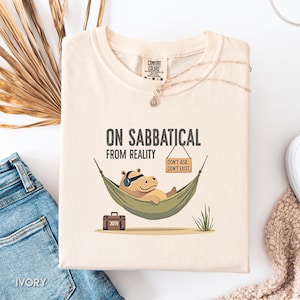 May include: Ivory-colored t-shirt with the text "ON SABBATICAL FROM REALITY." A cartoon hippo wearing headphones relaxes in a hammock. A sign reads "DON'T ASK. DON'T EXIST." A small suitcase is at the bottom. The shirt is folded on a surface.