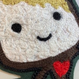 May include: Embroidered felt patch of a smiling cartoon character with blonde hair, brown eyes, and a red heart. The character is wearing a brown shirt and has a green background.