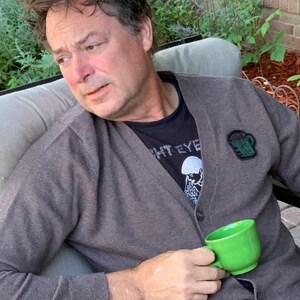 May include: A man wearing a brown cardigan with a black t-shirt underneath. The t-shirt has a white graphic of a person with a mohawk and the text "White Eye". The man is holding a green mug in his right hand.