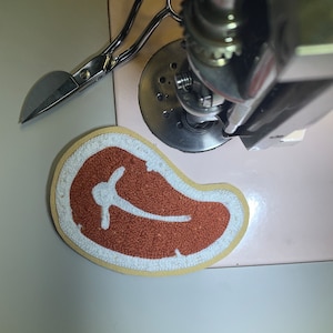 May include: A white and brown embroidered patch shaped like a steak. The patch is sewn onto a white surface.