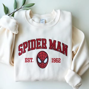 Może przedstawiać: Kremowa bluza Spider-Man z czerwonym haftowanym tekstem i grafiką maski Spider-Mana. Tekst brzmi "SPIDER MAN EST. 1962". Bluza ma okrągły dekolt i długie rękawy, złożone na mankietach.