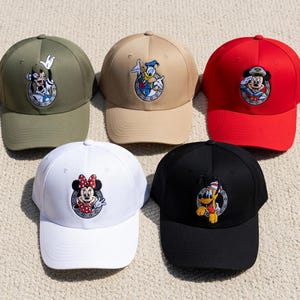 Puede incluir: Cinco gorras de béisbol en varios colores: verde oliva, beige, rojo, blanco y negro. Cada gorra presenta un personaje de dibujos animados diferente bordado en la parte delantera, dentro de un círculo.